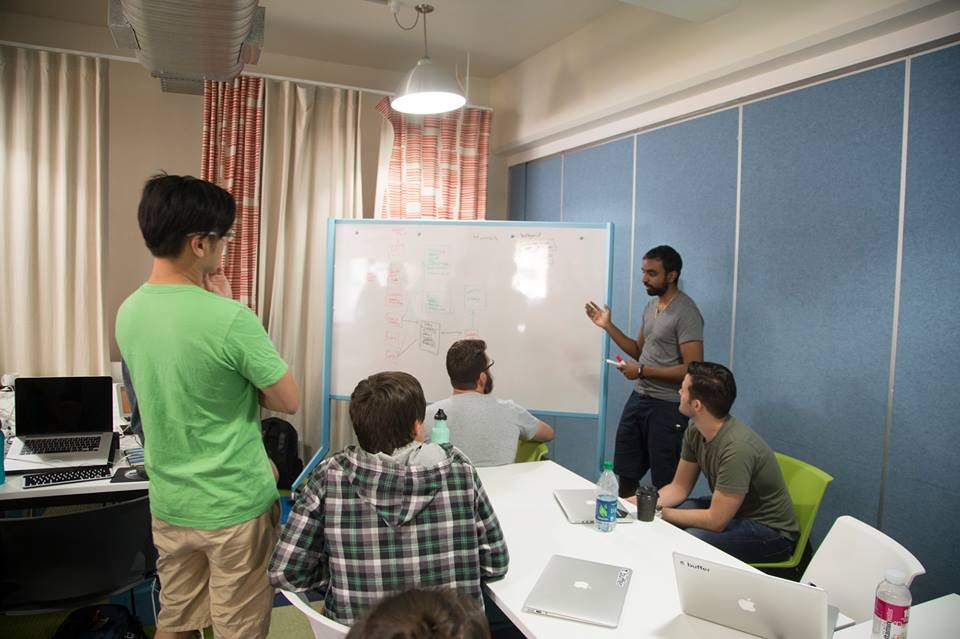 Sunil at a whiteboard covered in boxes and arrows, talking through a diagram with four teammates seated around a table.
